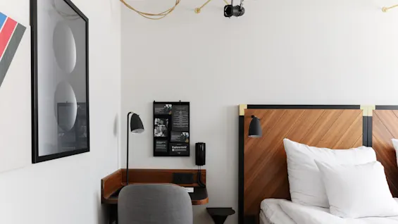 Ett modernt hotellrum med träpanel som sänggavel, vita sängkläder, ett skrivbord, stol, väggkonst och belysning.
