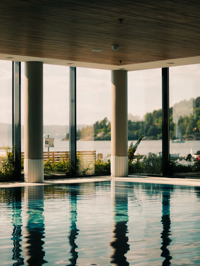 En inomhuspool med stora fönster som erbjuder en naturskön utsikt över en sjö omgiven av frodiga gröna kullar under en ljus himmel.