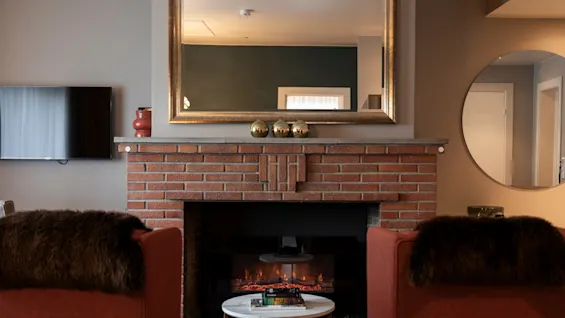 A cozy living area at Home Hotel Grand Bodø with a brick fireplace, two armchairs, and a large mirror.