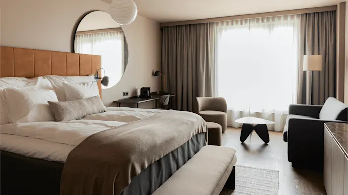 Et moderne hotellrom med en prydelig oppredd seng og et rundt speil, et lite skrivebord med en stol, samt en sittegruppe med lenestol, sofa og bord ved siden av store gardinerte vinduer.