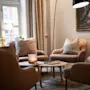 Fire beige lænestole omkranser et lavt bord med et skakspil og stearinlys, placeret i en hyggelig stue med et stort vindue, gardiner og kunst på væggen.