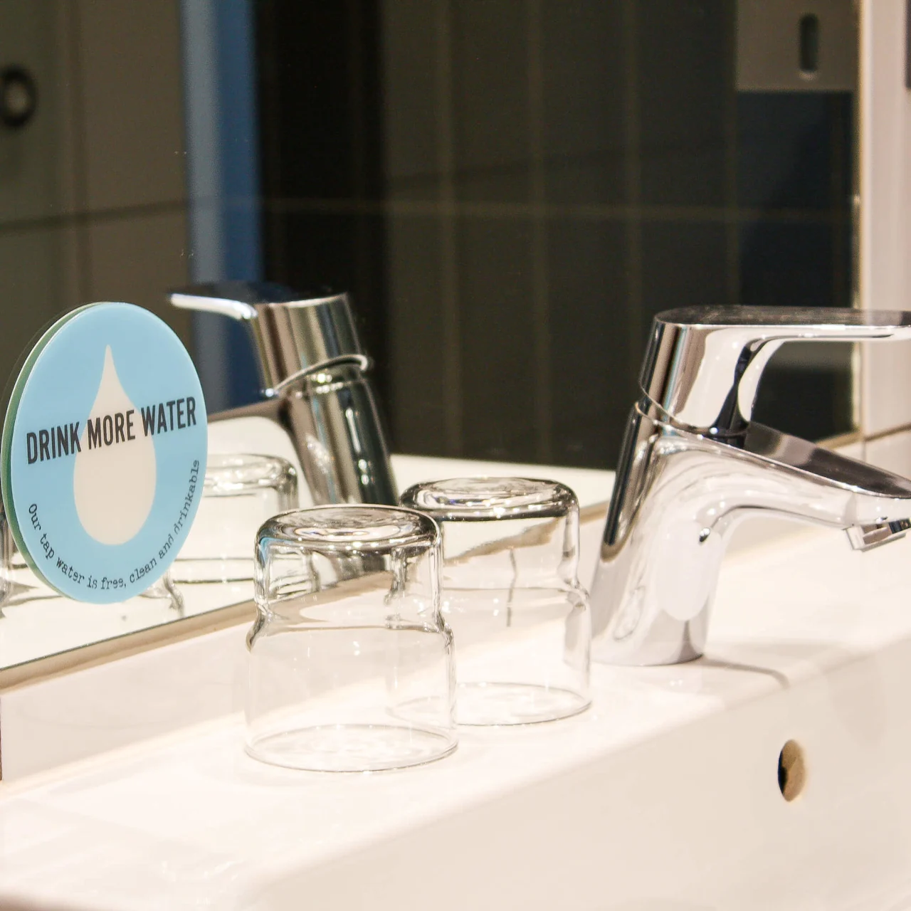 A faucet stands on a bathroom sink beside upside-down glasses, reflected in a mirror. A sign reads: “DRINK MORE WATER. Our tap water is free, clean and drinkable.”