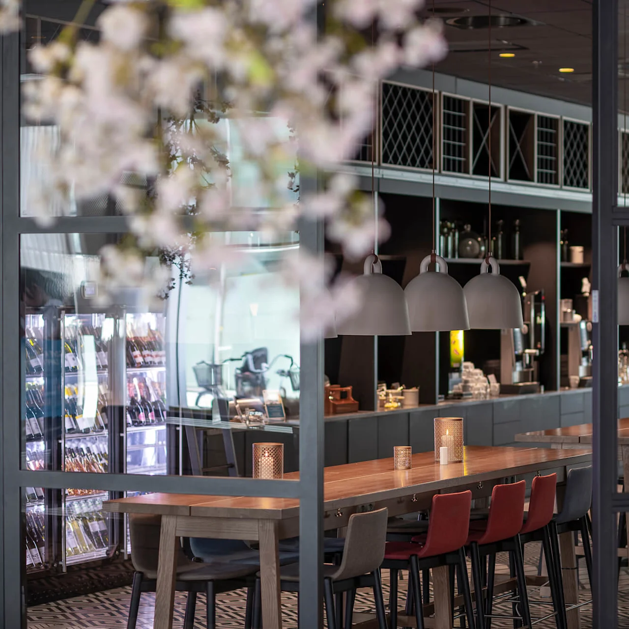 Long wooden table with red and black chairs in a modern cafe, illuminated by pendant lamps. Glass walls reveal fridges and shelves stocked with items in the background.