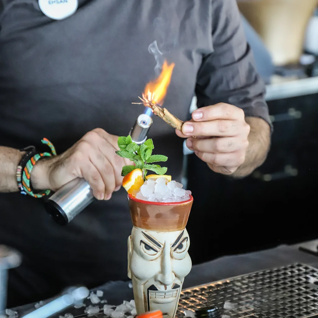 A person carefully torches a garnish over a vibrant tiki cocktail, enhancing its aroma and presentation. The name tag reads EHSAN.