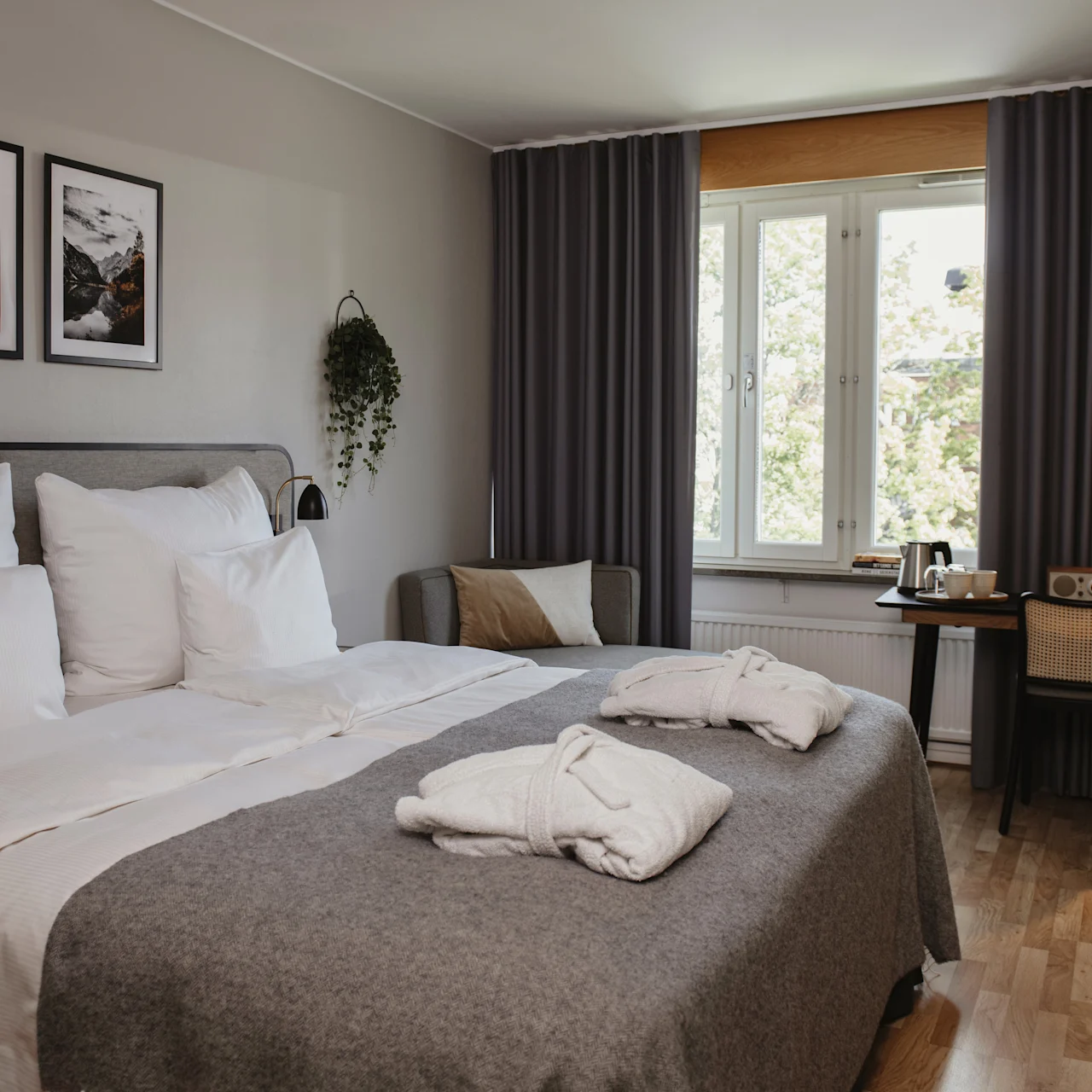 A neatly made double bed with two folded bathrobes sits in a hotel room. A desk with a lamp is by the curtained window, and framed art decorates the wall.