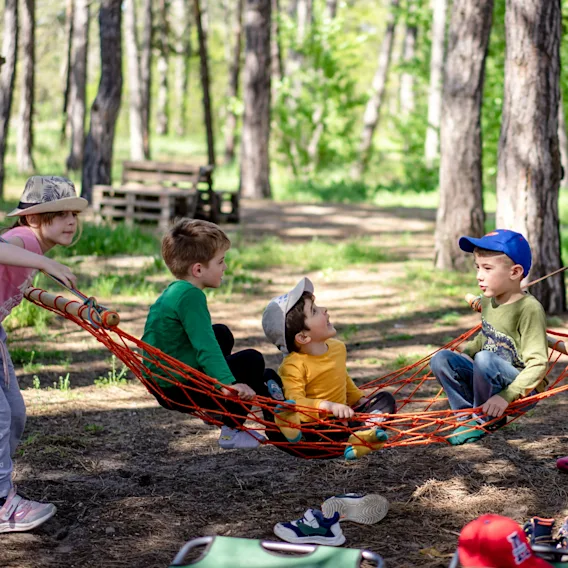 Barn leker i en hengekøye i en solrik skog. Ett barn dytter hengekøyen mens andre sitter og prater.