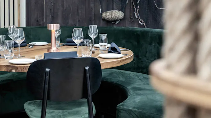 A round wooden table set with glasses, plates, and a copper lamp, surrounded by green velvet seating, in a rustic dining area with nets on dark wooden walls.