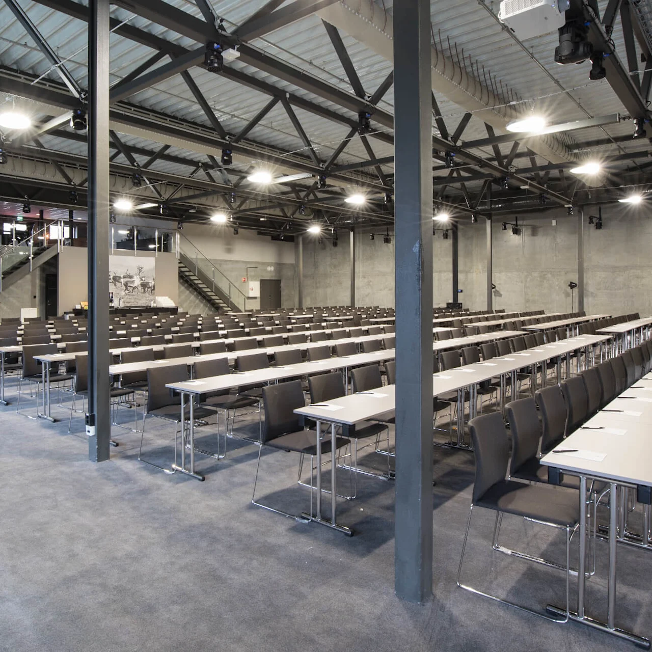 Rows of chairs and tables are set up for a conference in a large, industrial-style room with exposed beams. Stairs lead to an upper level. The space is well-lit.