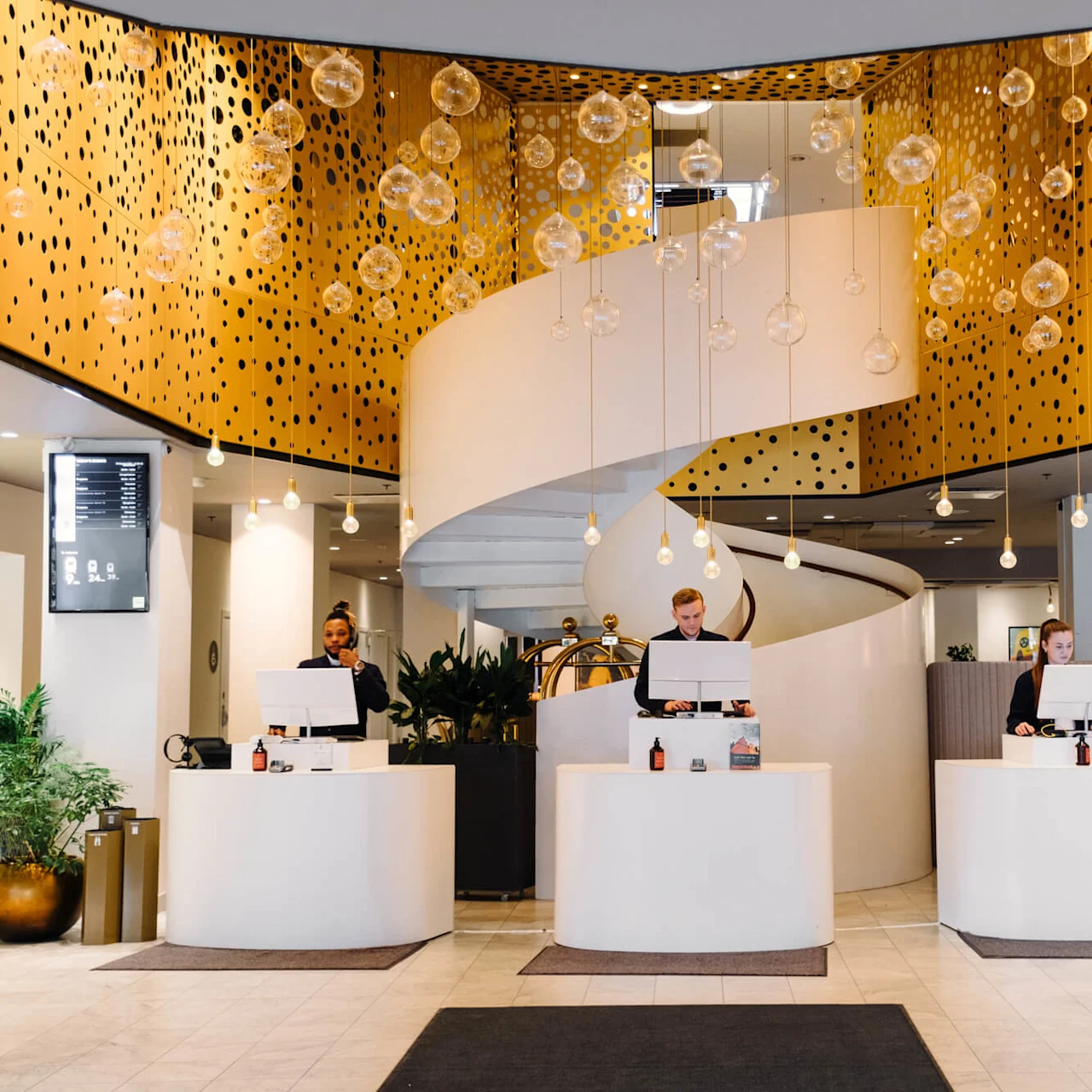 Reception area with three people behind modern, white desks under a decorative, golden ceiling with hanging glass orbs. A spiral staircase ascends in the background. Plants adorn the space.