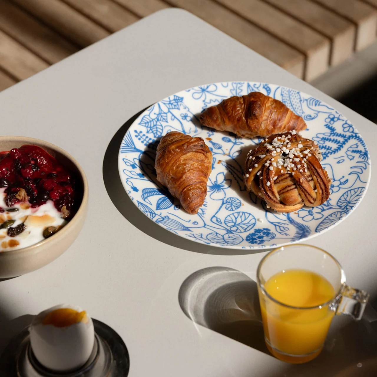 Croissants and a pastry rest on a patterned plate beside a glass of orange juice, yogurt with berries, and an egg, set on a table with wooden seating.