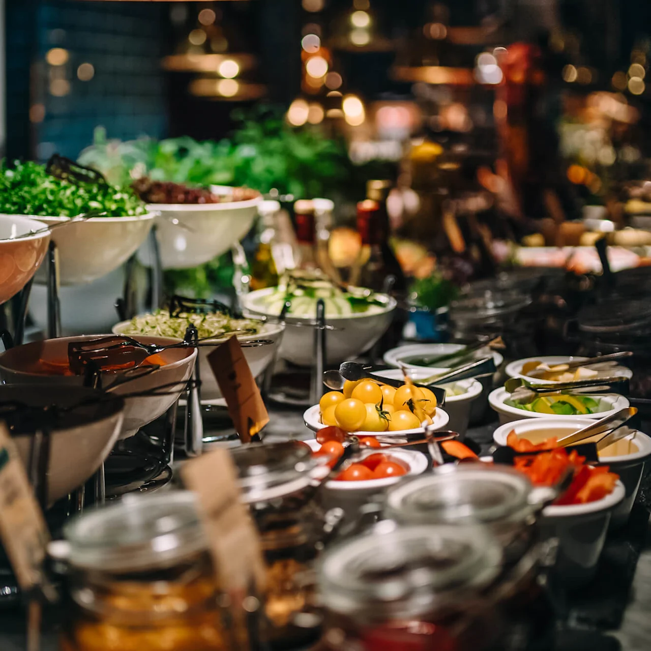 Bowls of fresh vegetables and salad ingredients are arranged on a buffet table, with leafy greens and colorful tomatoes, set in a dimly-lit dining area.