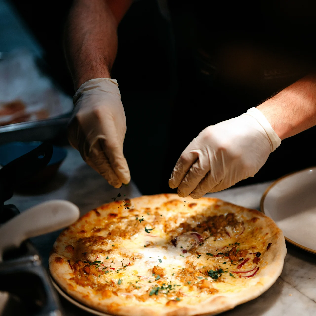 Pizza being garnished; chef’s gloved hands sprinkle herbs on a freshly baked pizza, set on a marble counter with other kitchen utensils around.