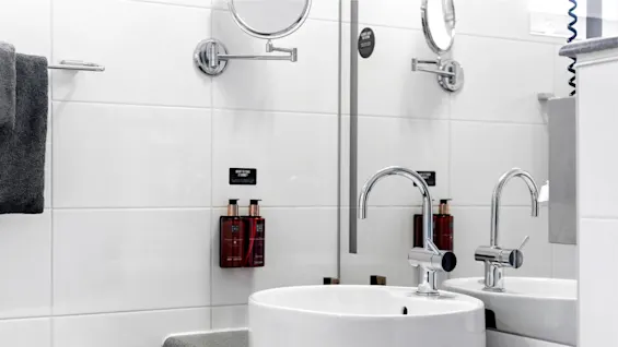 A modern bathroom sink with chrome faucets, a countertop, and white tiled walls. Accessories include a grey towel and brown soap dispensers.