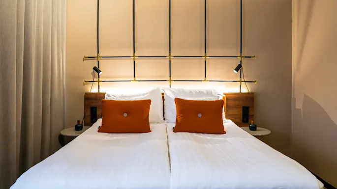 En pent oppredd seng med to oransje puter står mot en vegg utsmykket med geometrisk design og små, rettede lamper i et mykt opplyst, moderne rom.