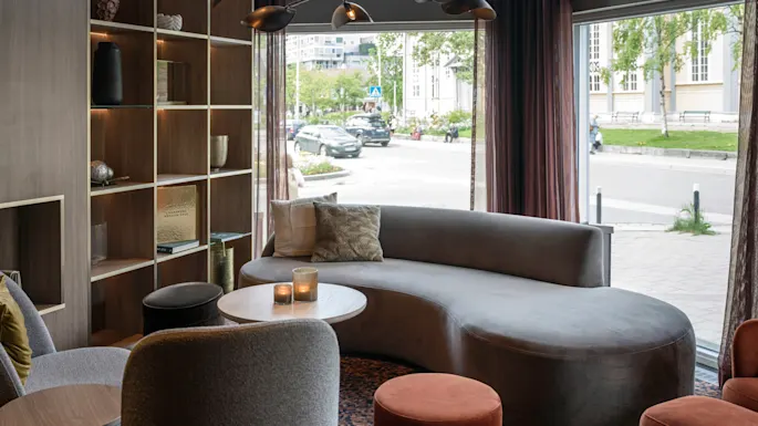 A curved gray sofa sits in a modern lounge, surrounded by chairs and a wooden shelf with decor items. Large windows reveal a street with trees and parked cars outside.