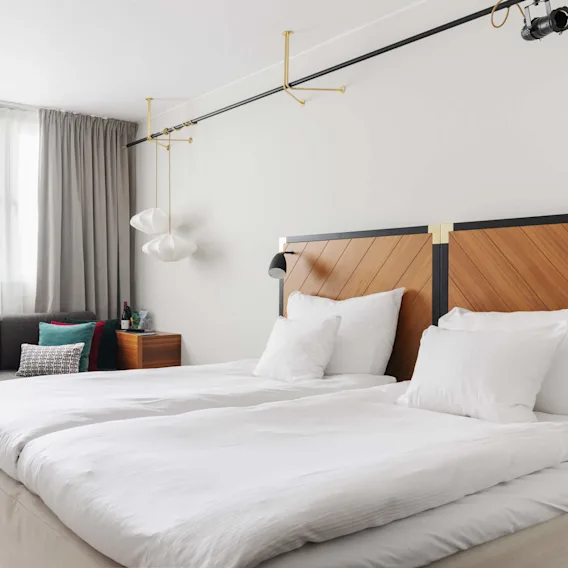 Moderne soveværelse med en pænt opredt seng, stilfulde lysarmaturer og et siddeområde med en grå sofa i et rent, lyst rum.