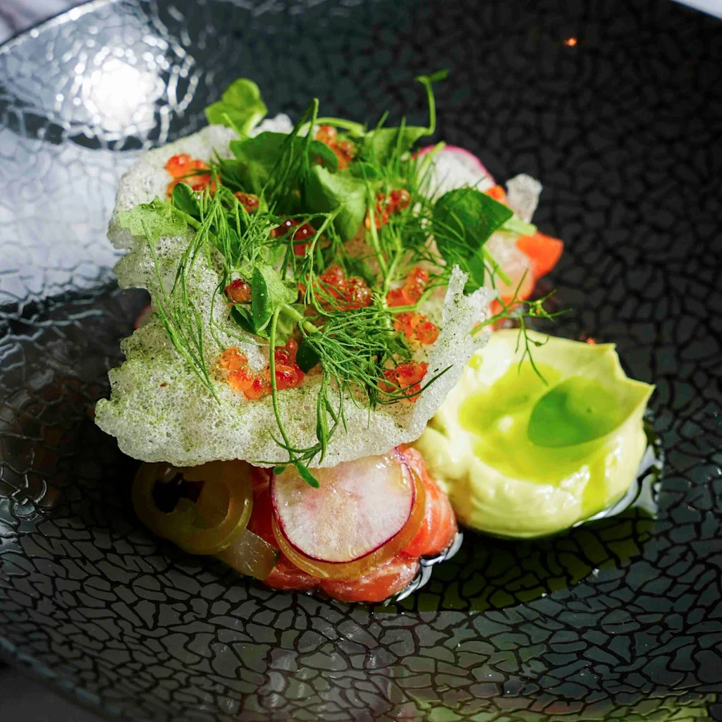 En gourmetrett med lagdelte ingredienser, toppet med friske urter, rogn og en kremet saus på en mørk tallerken.