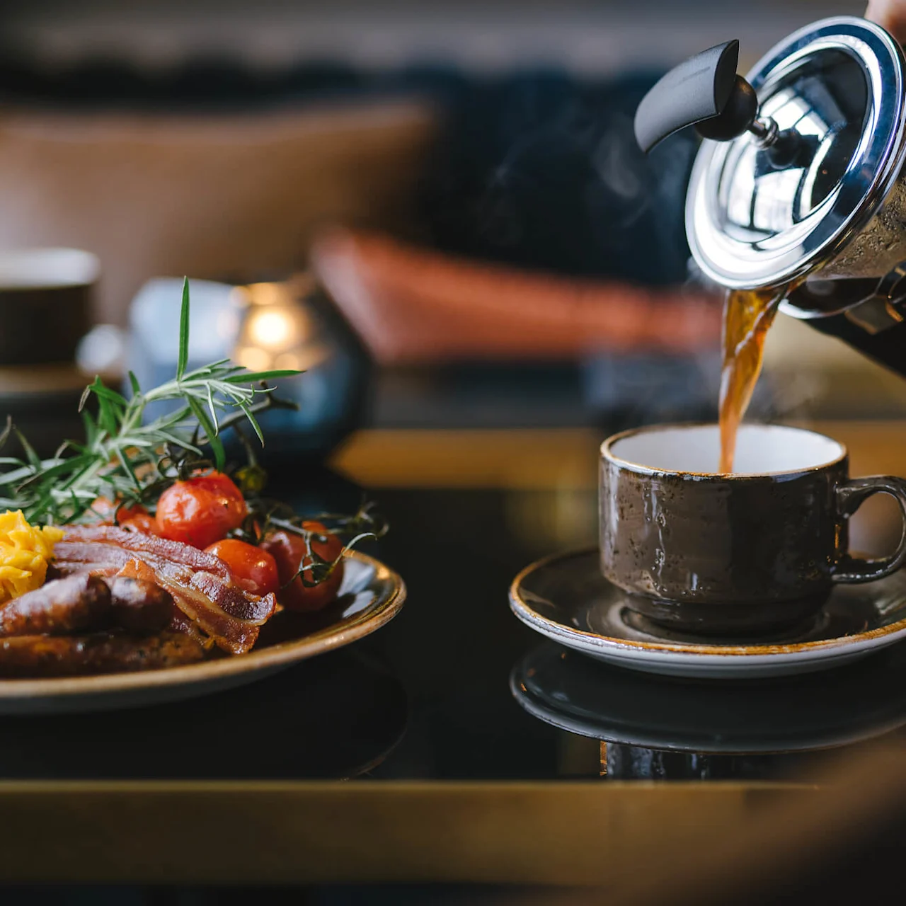 Coffee is being poured from a French press into a cup on a table, next to a plate of breakfast with bacon, scrambled eggs, cherry tomatoes, and herbs.