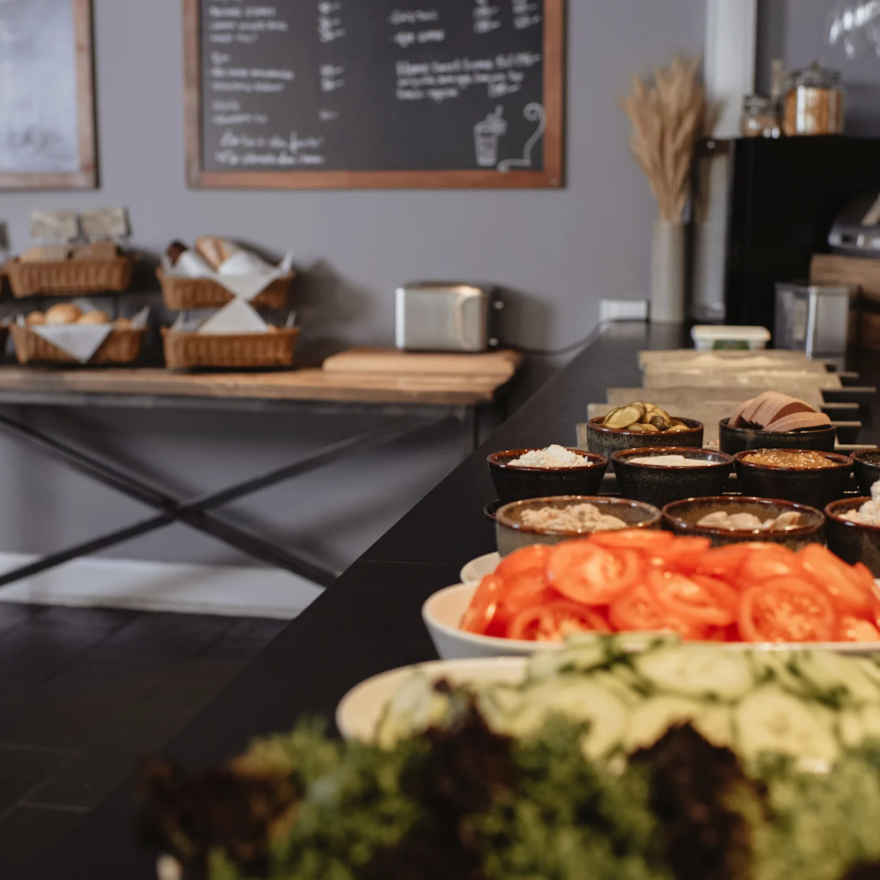 Salad ingredients are displayed in bowls on a counter; baskets of bread sit on a table in a café setting with chalkboard menus on the wall.