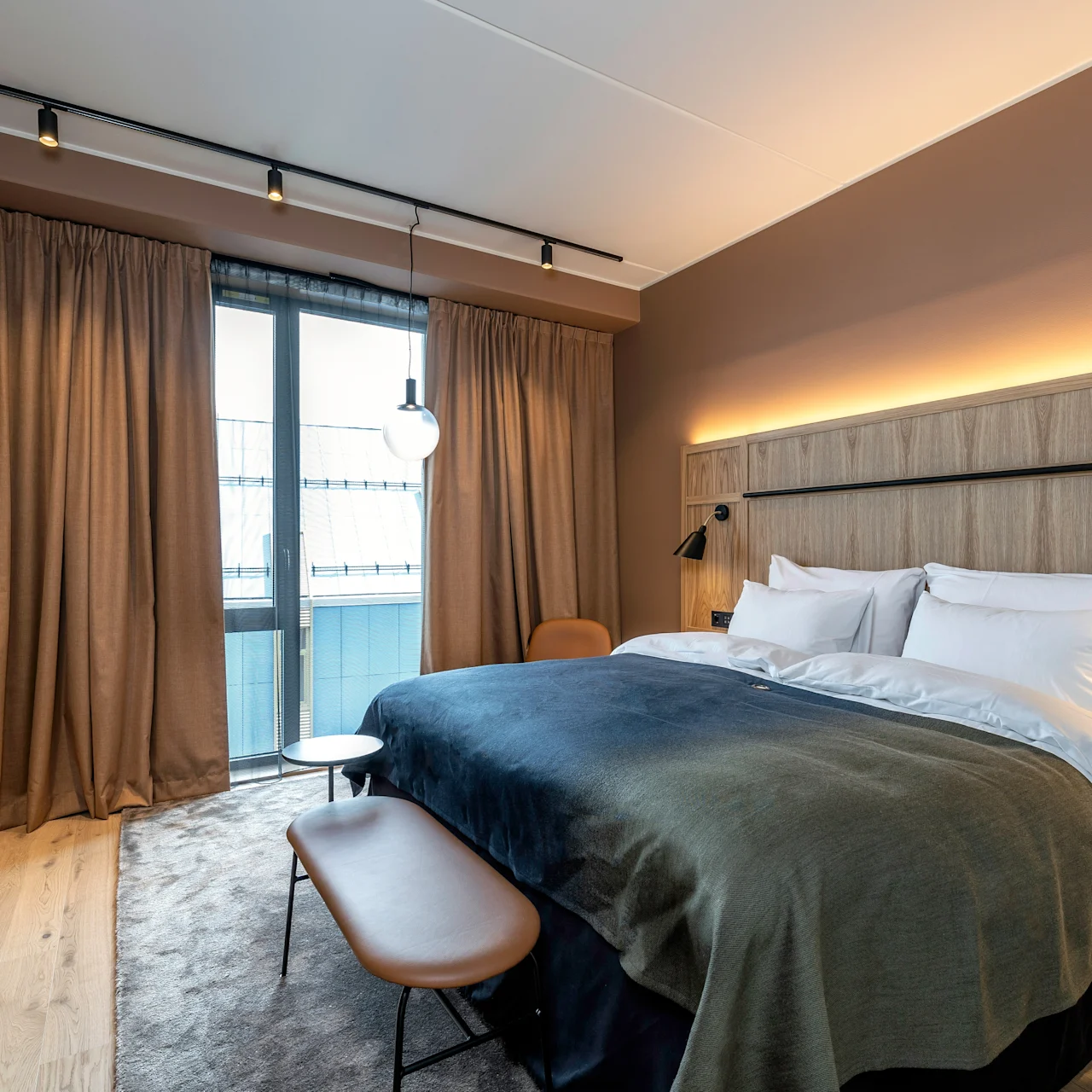En pænt redt seng med hvide puder og et mørkt sengetæppe, oplyst af væglamper, står mod træpanel i et moderne hotelværelse med gardiner og et stort vindue.