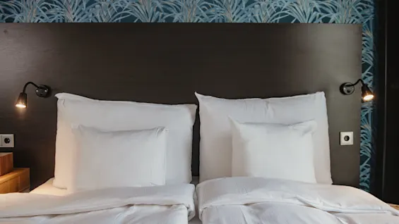 A bed with white linens and two pillows sits against a wooden headboard. Two small wall lamps are turned on, flanked by a blue patterned wallpaper.