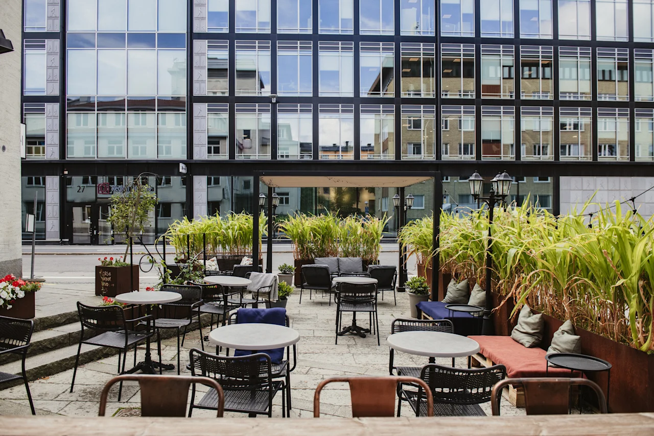Outdoor seating area at Home Hotel Uppsala with tables, chairs, and lush green plants, set against a modern building. Text: 27.