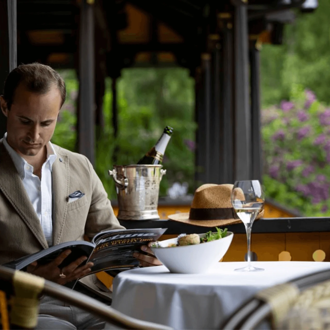 A man in a beige suit reads a magazine at a restaurant table outdoors. A bottle of champagne, hat, and glass of wine are on the table.