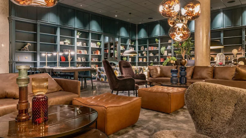 Luxurious leather sofas and armchairs surround a polished wooden coffee table in a library setting, with warm pendant lighting overhead. Books and decorative objects fill the shelves along the walls.
