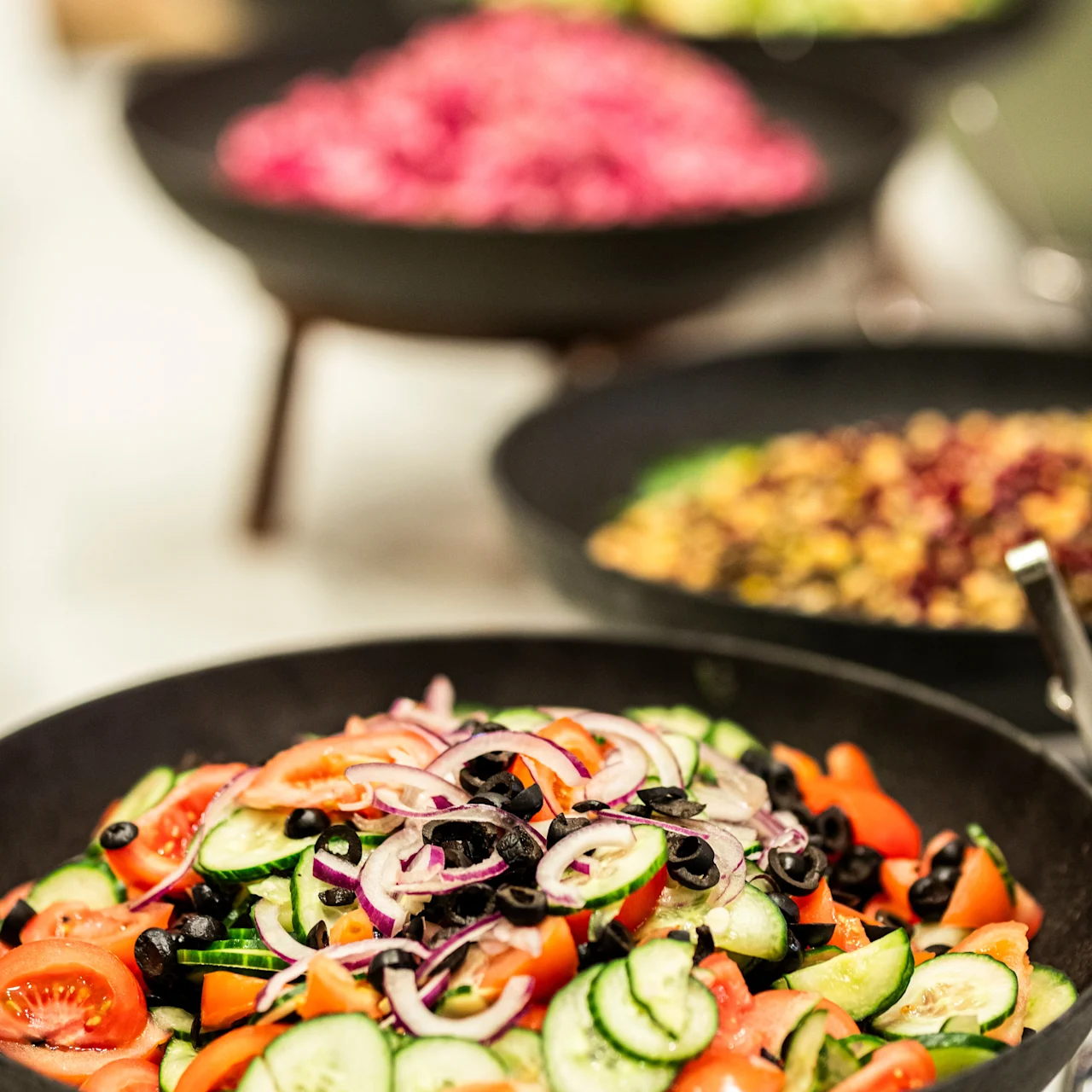 A bowl of fresh salad with cucumbers, tomatoes, olives, and red onions is placed on a buffet table, surrounded by other colorful salad dishes.