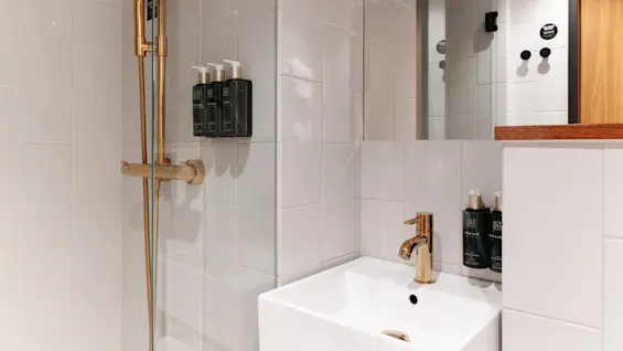 En modernt badrumshörn med en guldfärgad dusch och ett vitt handfat, hygienartiklar på hyllor, reflekterande i en glasduschdörr.
