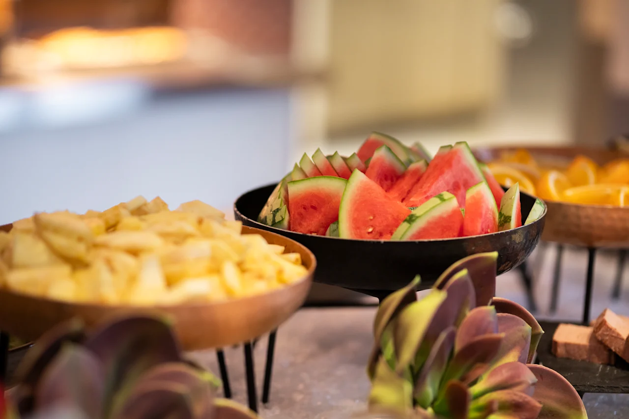 Eloisa esillepano tuoreita hedelmiä, mukaan lukien viipaloitu vesimeloni ja muita vaaleita hedelmiä, järjestettynä kulhoihin buffetissa.