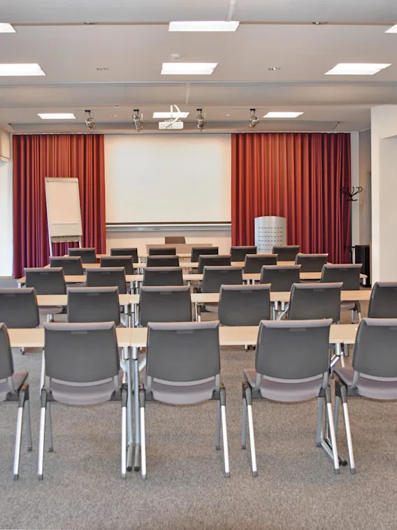Rader av tomma stolar och skrivbord vända mot en vit skärm i ett väl upplyst konferensrum med röda gardiner, blädderblock och väggmonterad TV. En klädhängare och ett litet bord syns.