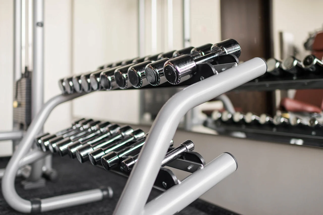 Dumbbells rest on a metallic rack in a gym, reflecting light from nearby mirrors, with exercise equipment surrounding the area.
