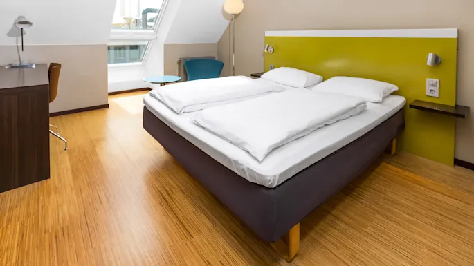 A double bed with white linens sits in a modern, bright room. A wooden desk with a chair is nearby, alongside a window, blue chair, and floor lamp.