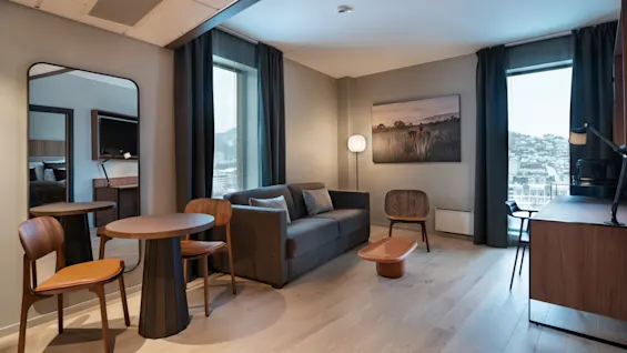 En moderne stue har en grå sofa med puter, et lite rundt bord med stoler og en gulvlampe. Store vinduer og et veggmaleri skaper en koselig atmosfære.