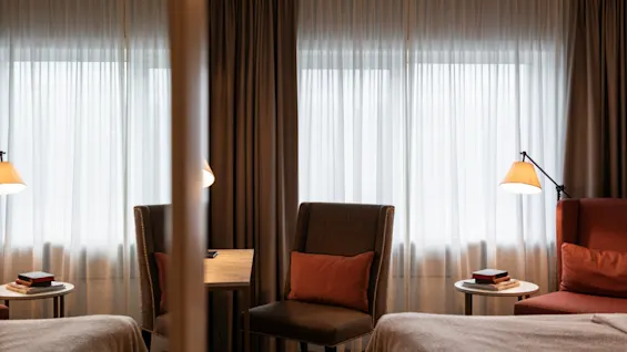 A comfortable hotel room at Home Hotel Grand Bodø with two beds, chairs, and a table, bathed in soft light from large windows.