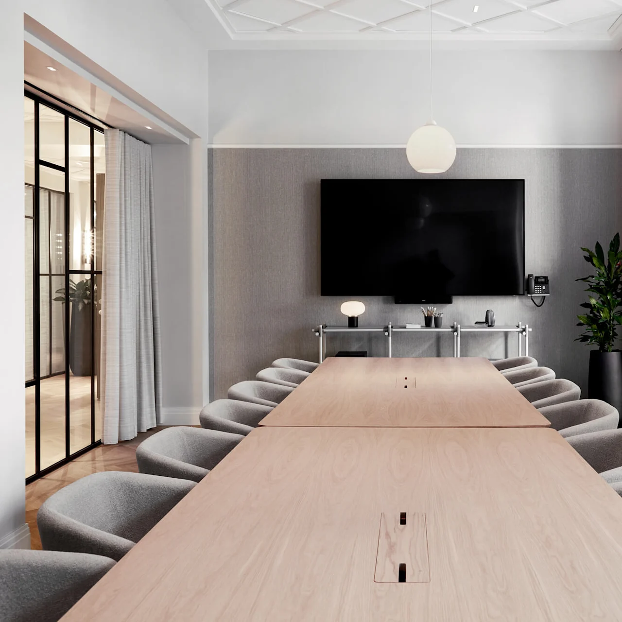 A long wooden conference table is surrounded by gray chairs in a room with a large screen, modern lighting, and potted plant, creating a professional atmosphere.