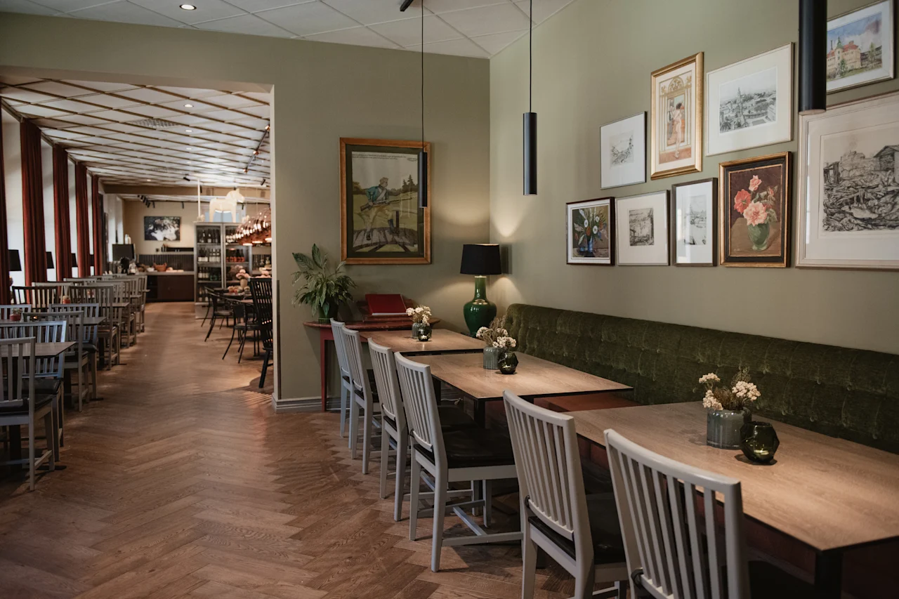 Et koselig restaurantinteriør med trebord, hvite stoler og en lang grønn fløyelsbenk. Innrammet kunst pryder den olivengrønne veggen.