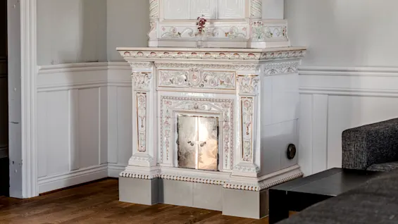 Elegant ceramic fireplace adorned with intricate floral patterns stands in a room, reflecting light from a partially open door, surrounded by white wainscoting and wooden floors.
