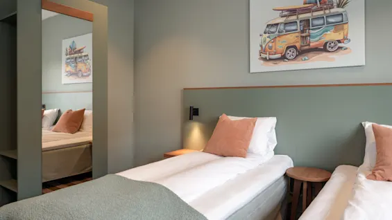 Two single beds with green blankets and peach pillows are beside a wooden stool in a room. A mirror reflects the beds, and a painting of a van decorates the wall.