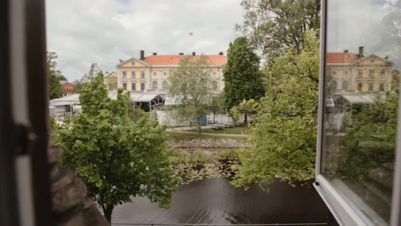 Ett fönster öppnar sig mot en vy av en flod med näckrosor, omgiven av gröna träd och historiska byggnader med röda tak i fjärran under en molnig himmel.