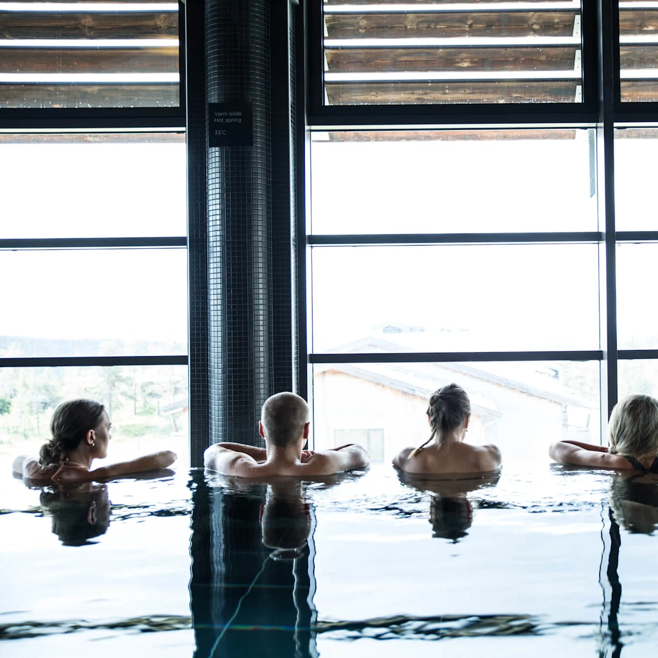 Five people relax in an indoor pool, looking out through large windows at a natural landscape. A sign reads "Varm kilde hot spring 35°C".