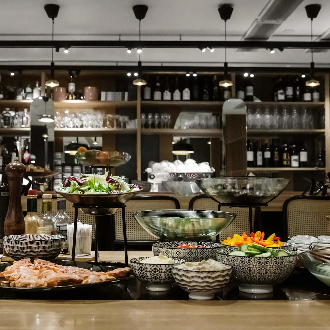 Et udvalg af retter og tilbehør er anrettet på et bord, hvilket antyder en buffetopsætning, i et rum med hyldedisplay af flasker og glas.
