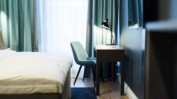 A bed with white sheets is positioned next to a desk and chair. A lit lamp sits on the desk, with teal curtains in the background.