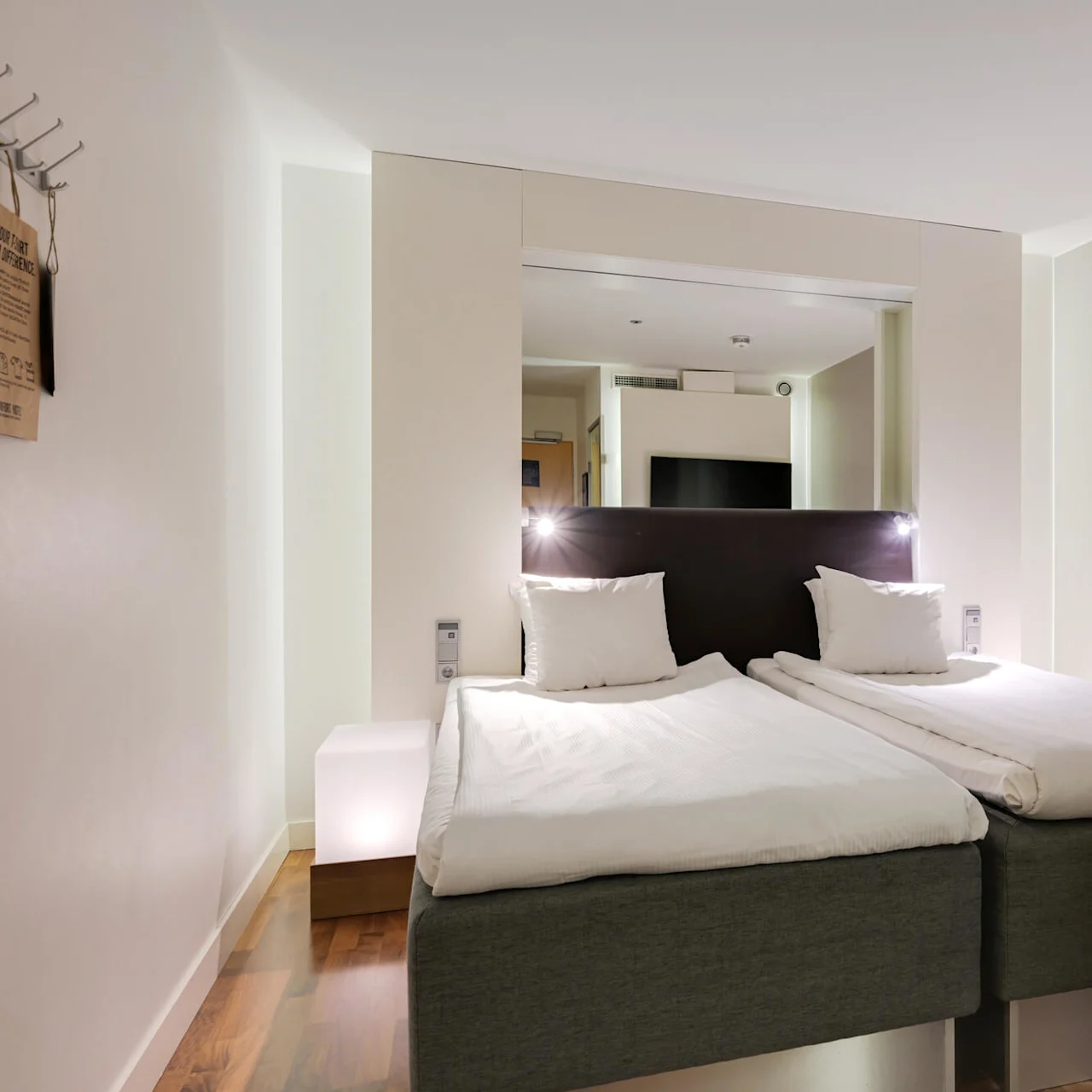 Två prydligt bäddade enkelsängar står bredvid varandra i ett modernt hotellrum. Ett trägolv och mjuk belysning skapar en varm, inbjudande atmosfär.