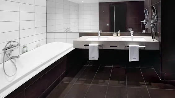 A modern bathroom with a white bathtub, dual sinks, and wall-mounted mirrors. Tiled walls and floor, with towels and toiletries arranged neatly.
