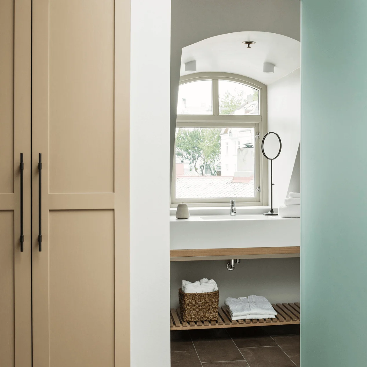 A bathroom with a large window above a white sink, wooden shelves below holding folded towels. Beige cabinets and a partial green partition are visible.