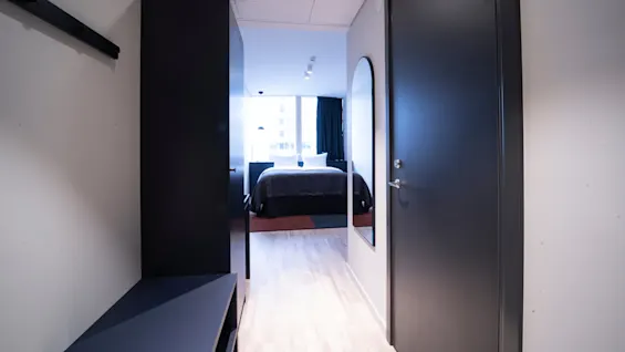 A hallway leads to a bright bedroom with a bed covered in dark linens. A large window illuminates the room, flanked by dark curtains. A wall mirror is on the right.
