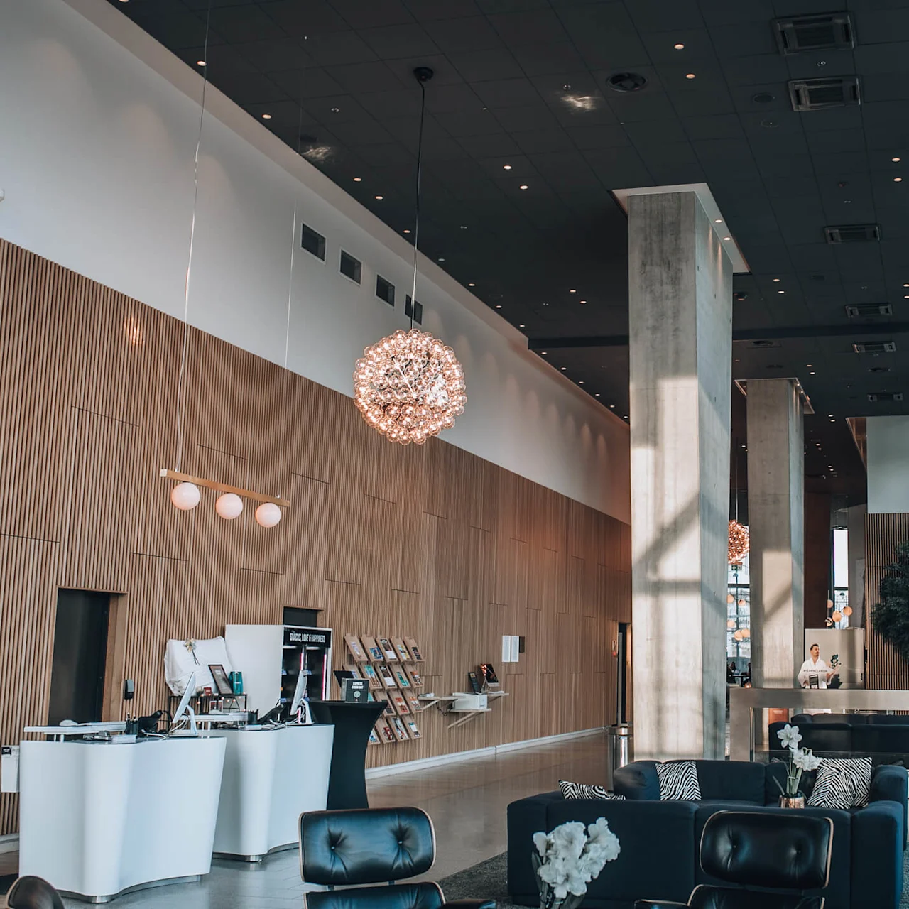 Rummelig hotellobby med moderne sorte lædersæder, en central træreception og store dekorative lysarmaturer. Gulv-til-loft-vinduer giver masser af naturligt lys.
