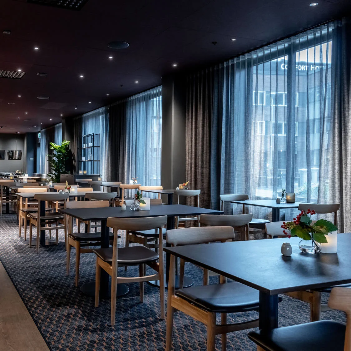 Restaurentbord, pent arrangert med lys og blomster, står under hengende lamper i et moderne, svakt opplyst spiseområde med store vinduer og mønstret teppe. Et "Comfort Hotel"-skilt er synlig utenfor.